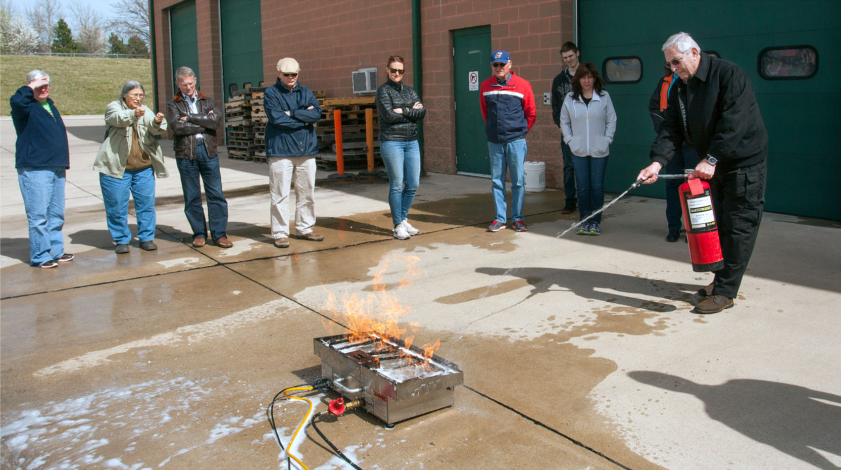 Man hold fire extinguisher to put out gas grill fire in fire safety training while group watches in background