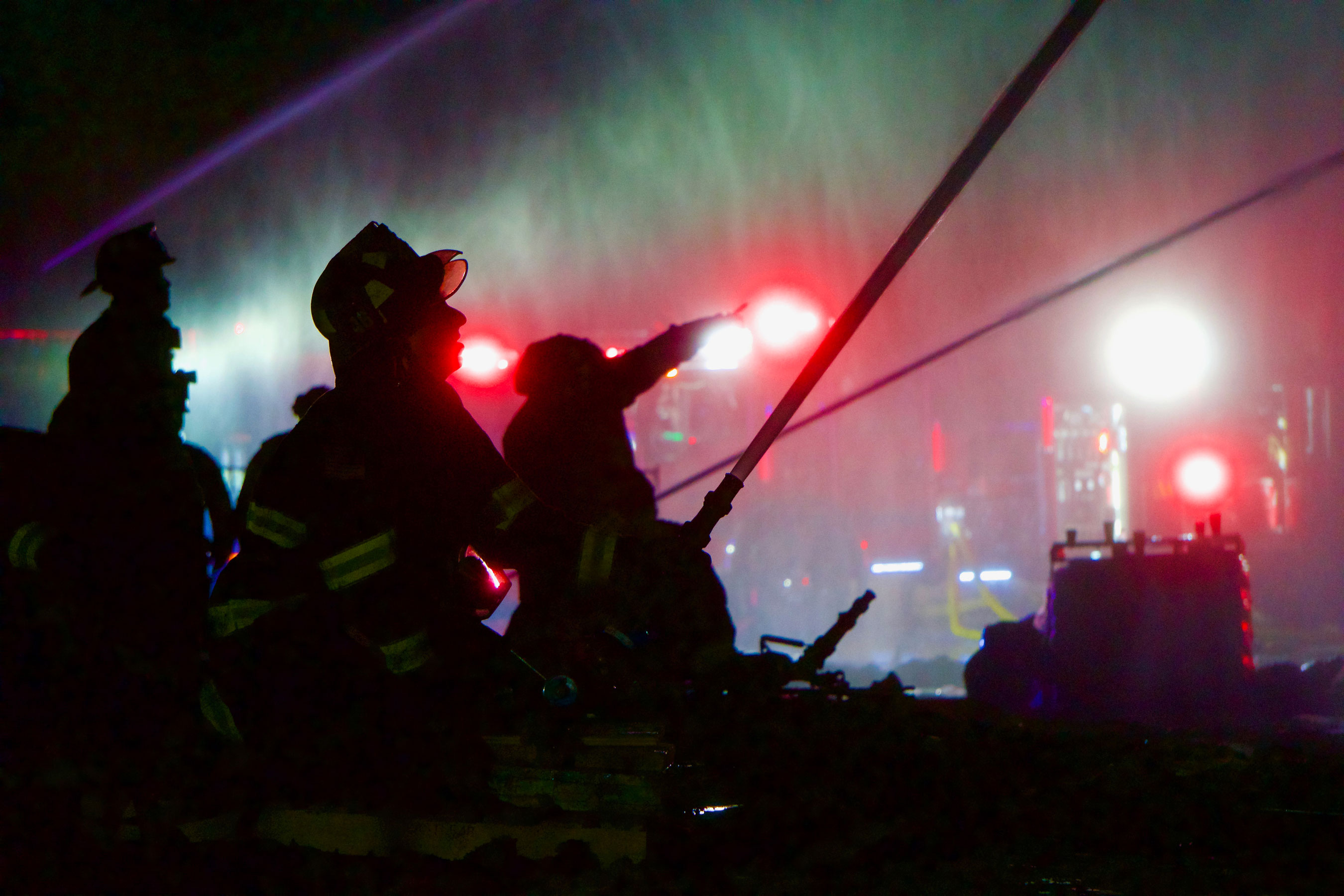 Silhouette of three firefighters and hose jets fighting fire at night with fire engine lights in background