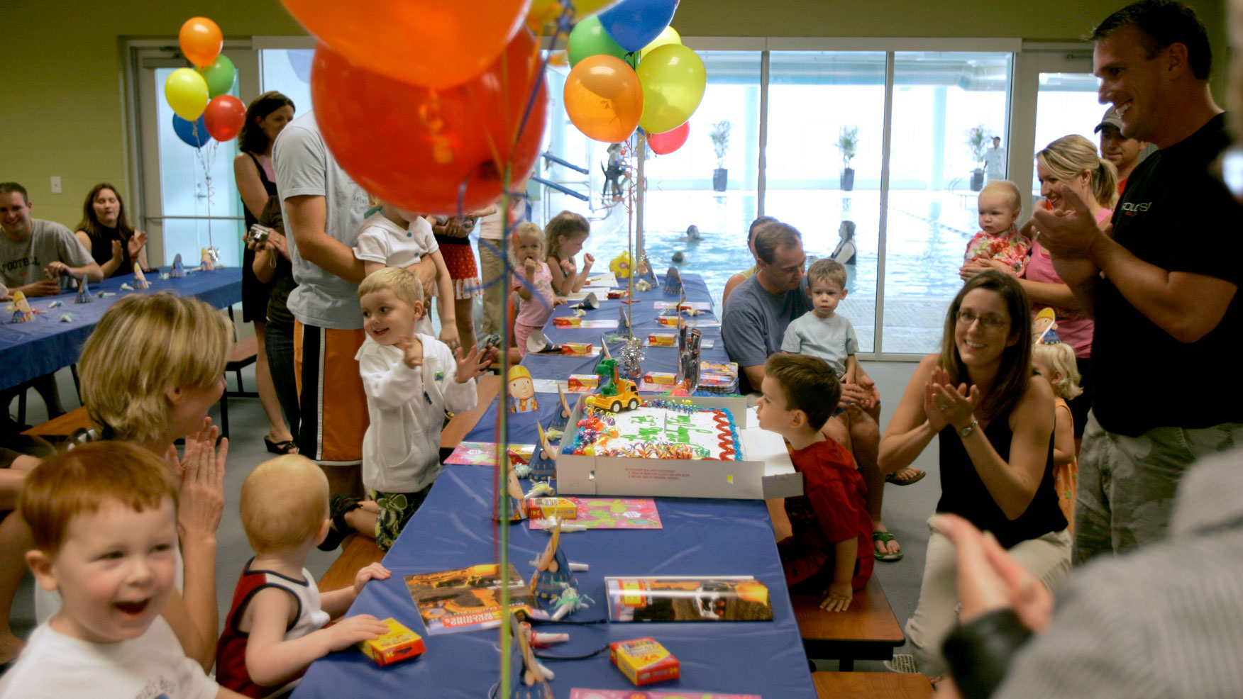 children and parents clap and smile during birthday party in party room next to pool with balloons