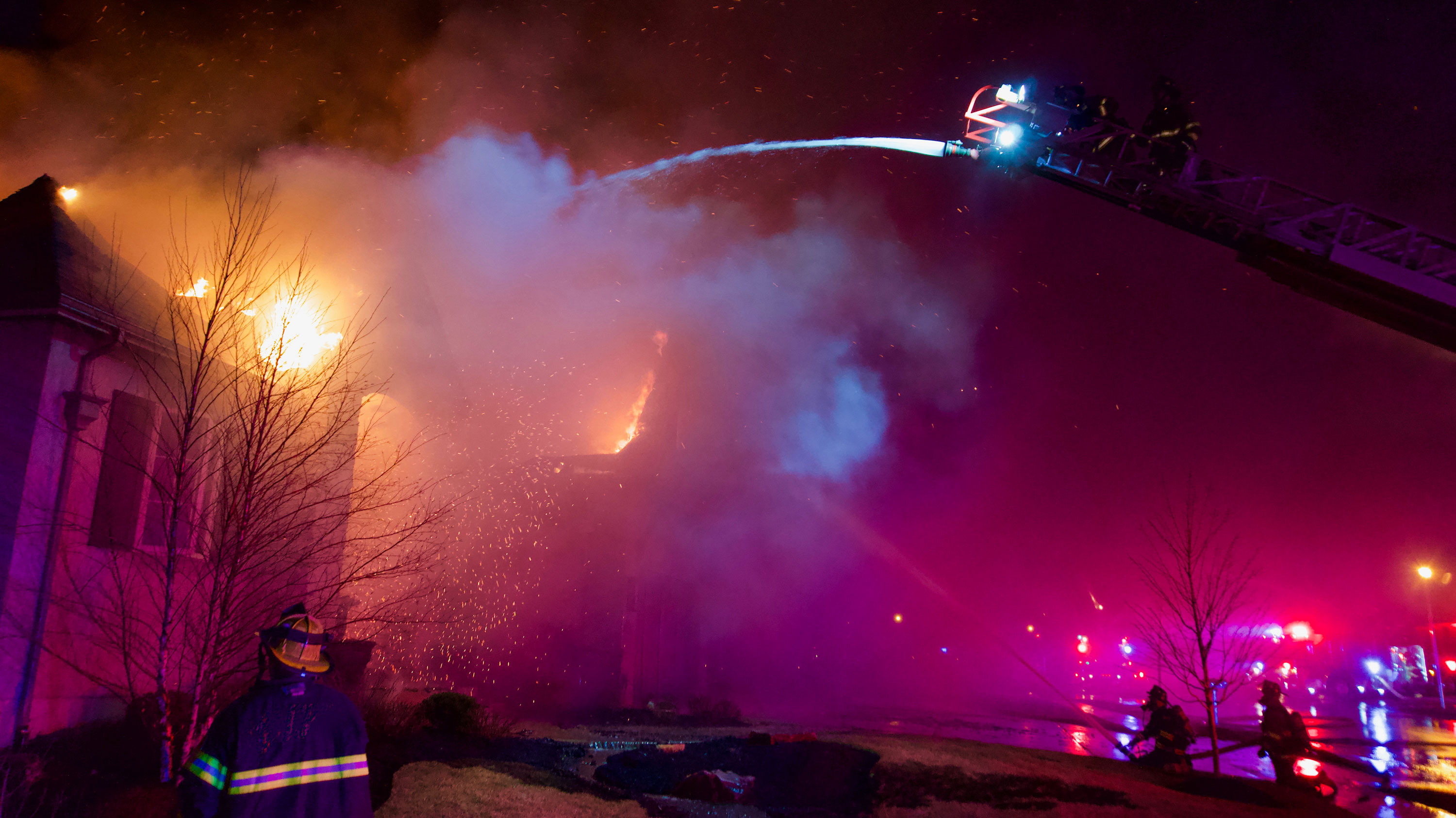Firefighters at top of ladder fight house burning from roof fire with aerial stance at night