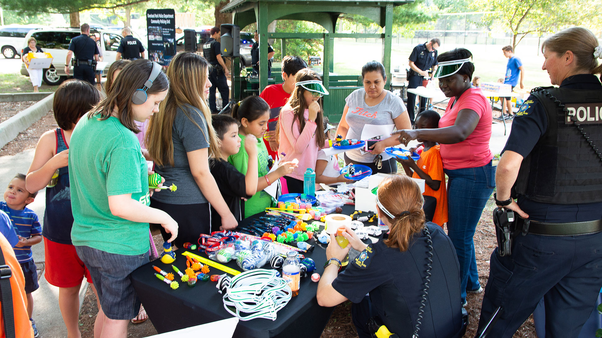 Group of children, adults, and police officers gathered around a table of toys and marketing merchandise at an event at an outdoor park