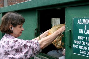 woman dumps recyclables in large green recycling dumpster while wearing headphones
