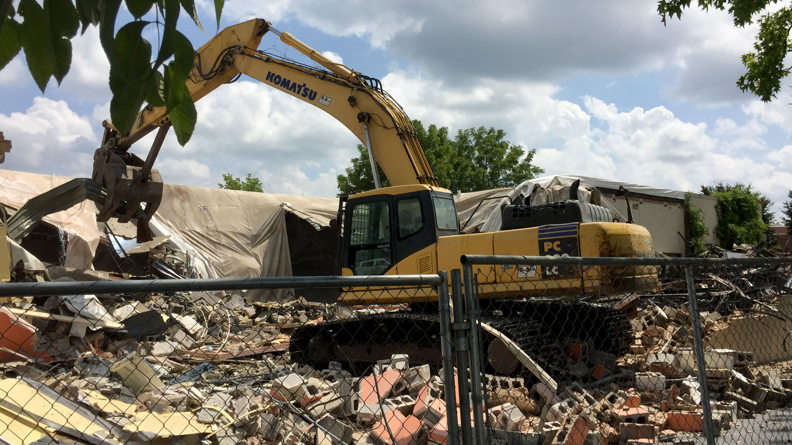 Yellow excavator knocks down building wall behind construction safety fence