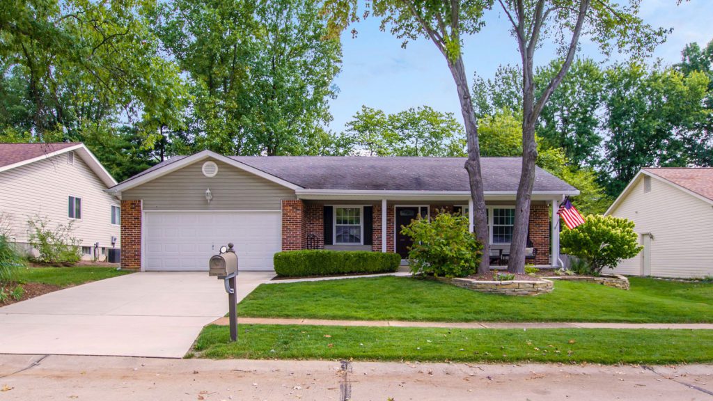Brick home with two-car garage, large trees, American flag and driveway on residential street