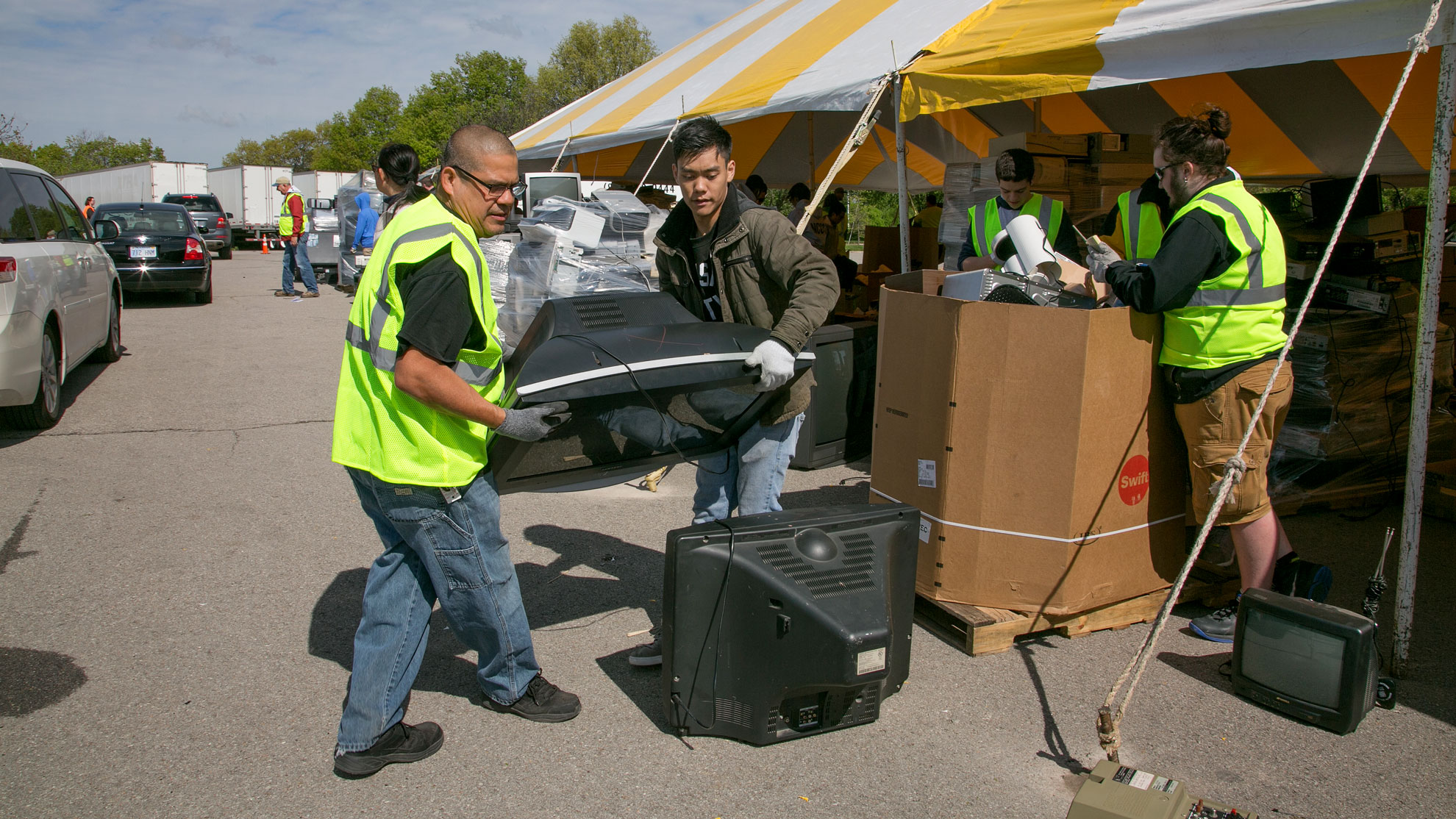 Two volunteers move television from vehicle to stack of electronics for recycling under large outdoor event tent
