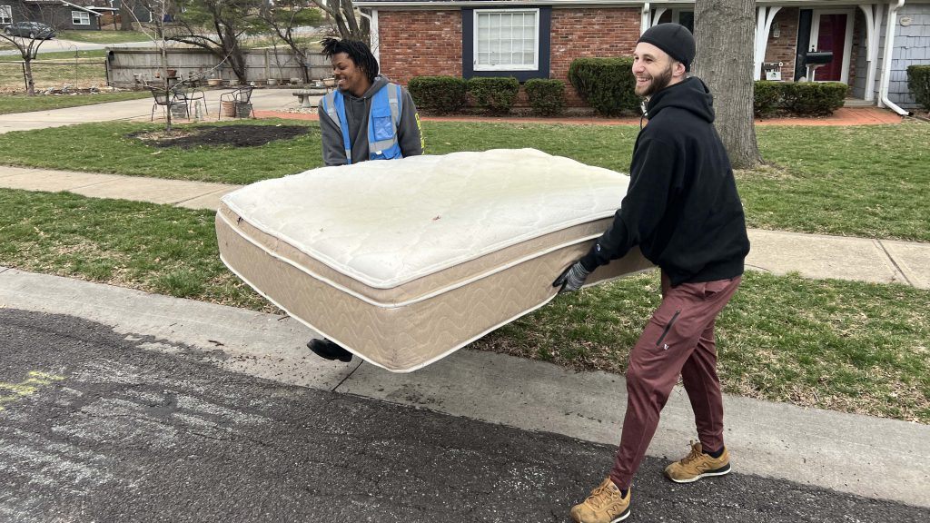 Two people work together to carry a mattress away from a house.