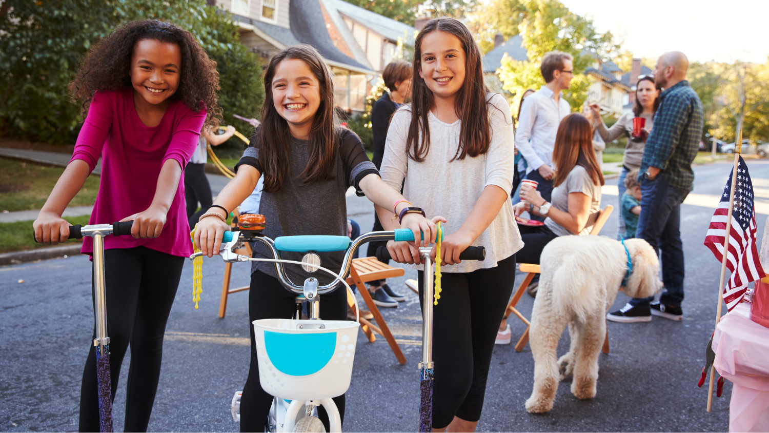 Three young girls on bikes and scooters smile for a photo while adults talk in a group in the background.