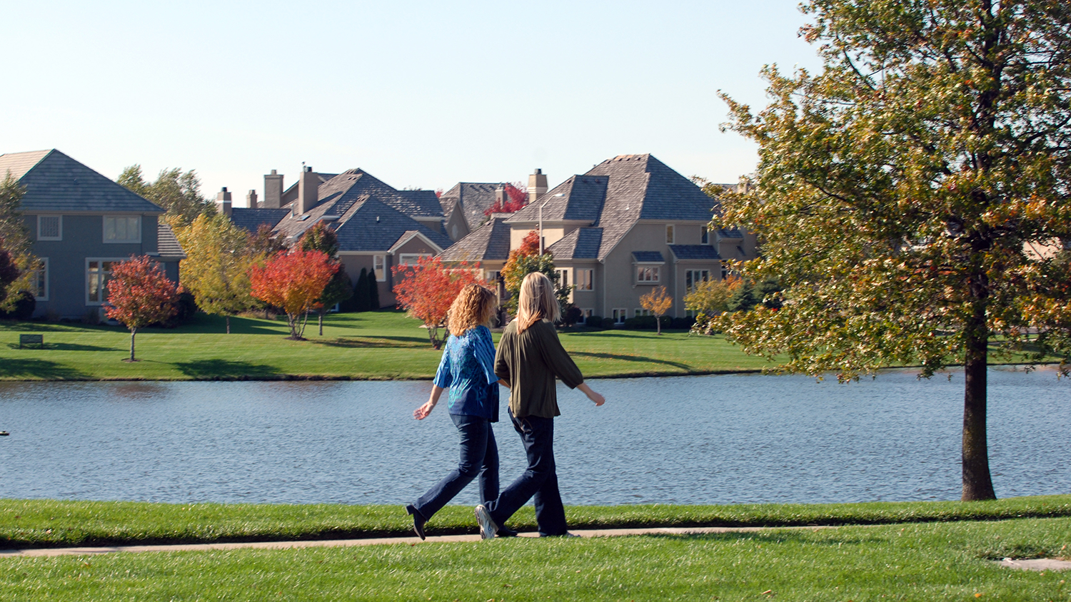 Two women walk side-by-side down a park trail with neighborhood homes visible across a lake.