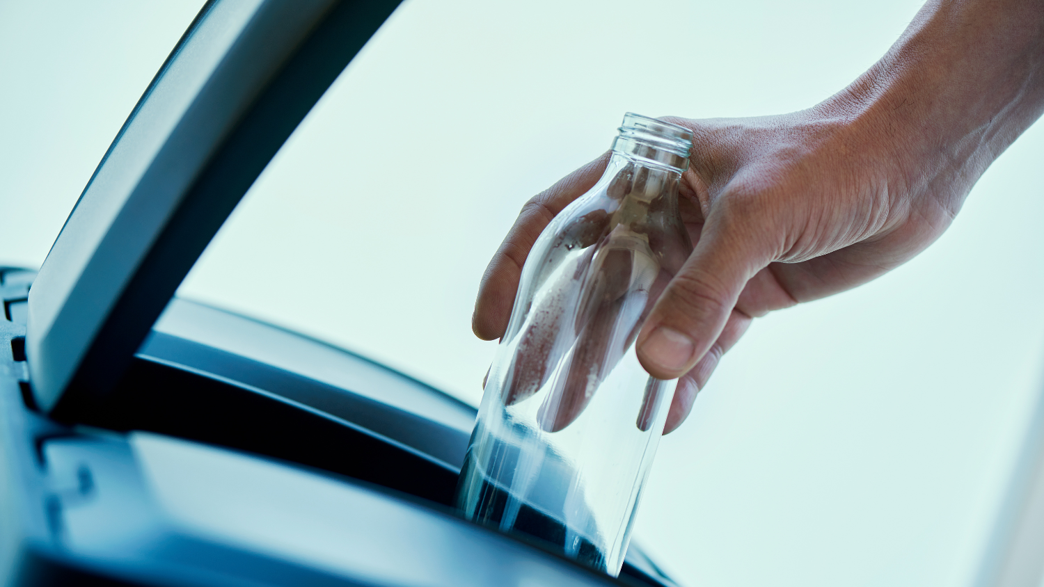 A close-up of a hand placing a glass bottle into a recycling can.