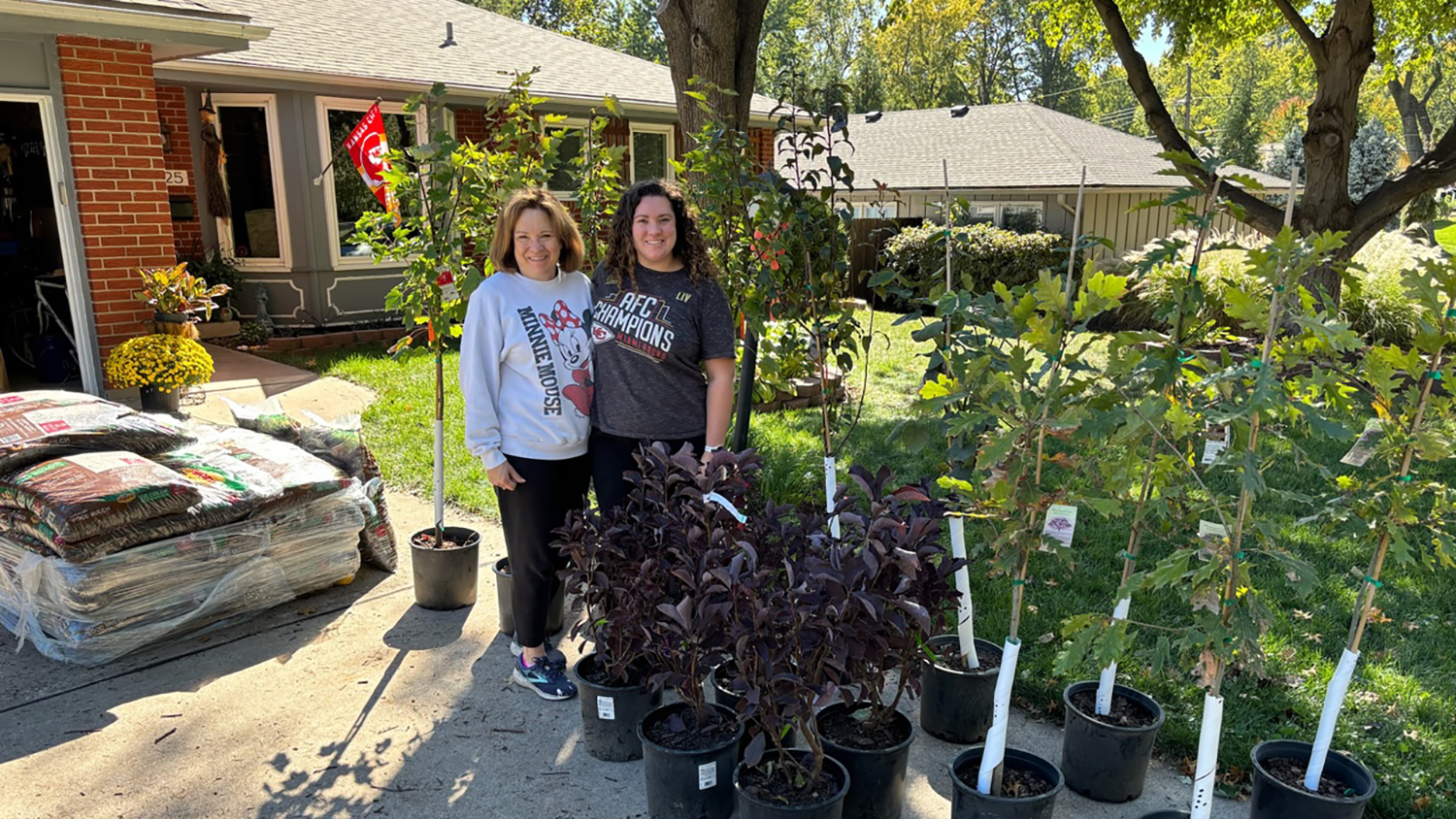 Two neighbors smile and pose next to several potted plants and trees sitting on a driveway.
