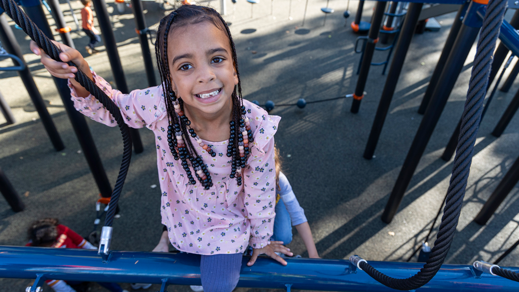 A young girl smiles at the camera while climbing on the playground at Strang Park.