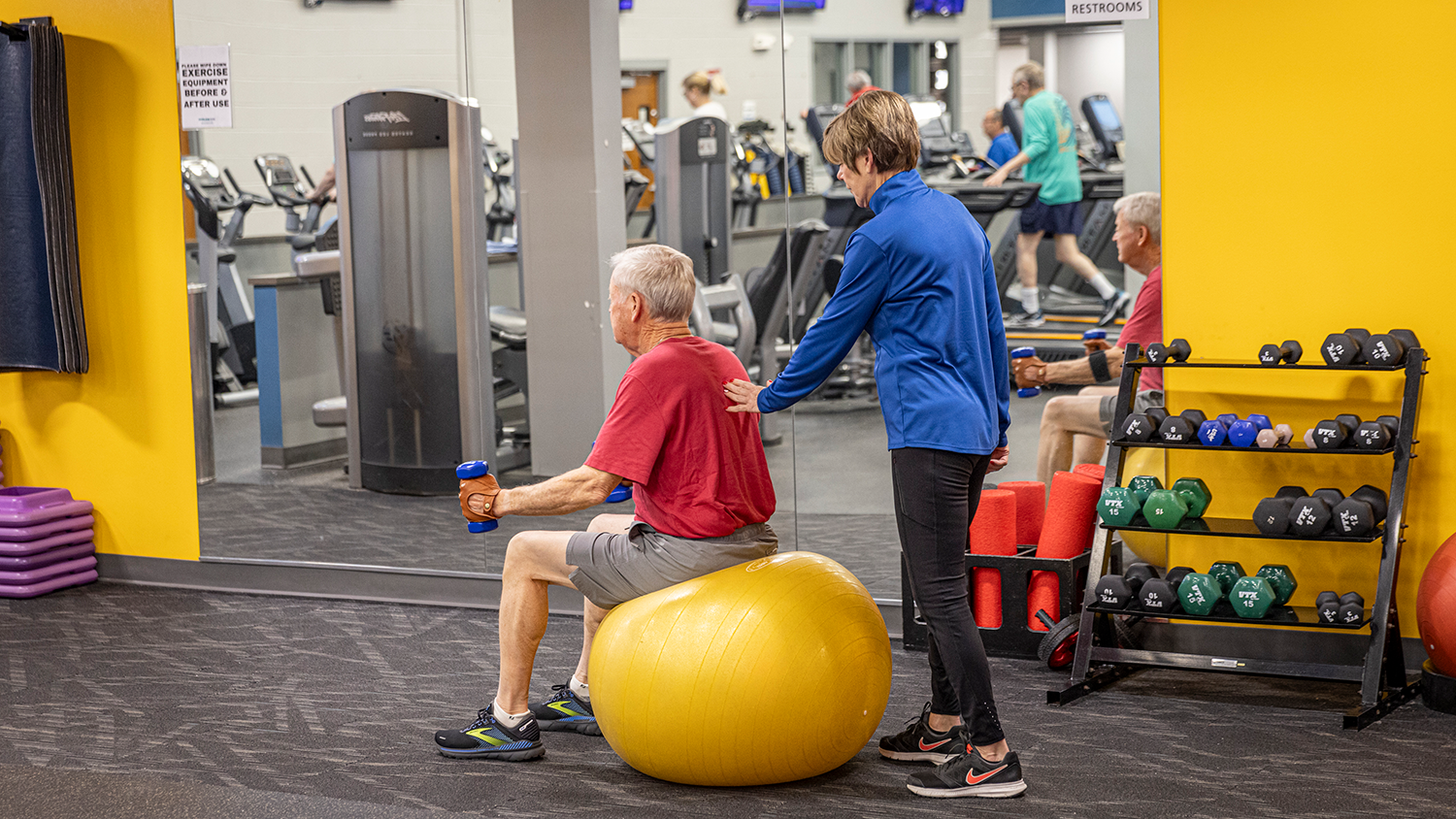 A man lifts weights while seated on a yoga ball at the fitness center. A friend supports behind him.