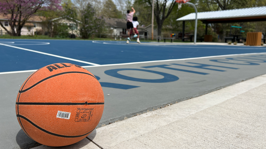 A basketball rests on the side of the outdoor basketball courts at Cherokee Park while two teenagers play in the background.