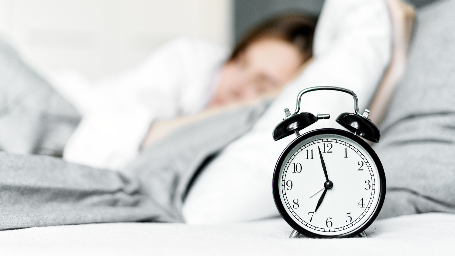 An alarm clock reads 6:58 as a woman sleeps in her bed.