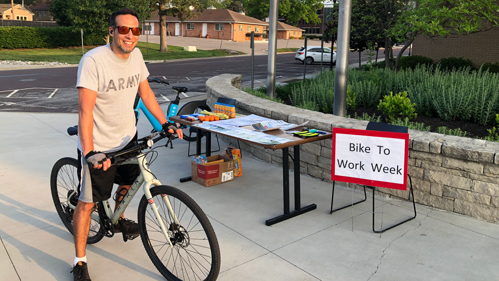 A man smiles on his bike, standing next to a table and sign that reads "Bike To Work Week."