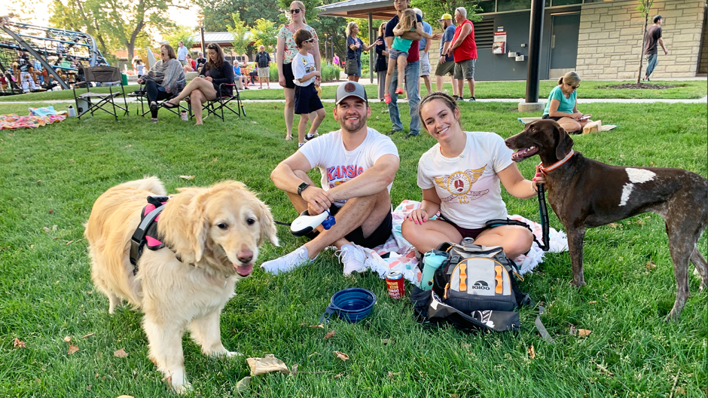 A couple and their two dogs sit on a blanket in a park.