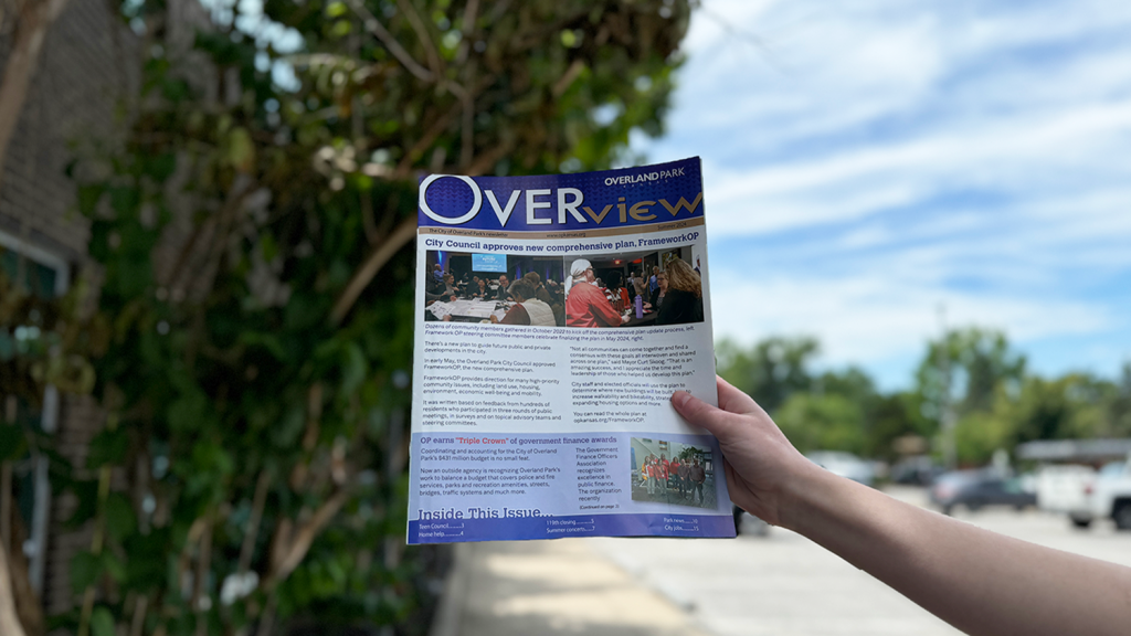 A hand holds Overland Park's summer 2024 Overview newsletter in front of City Hall.