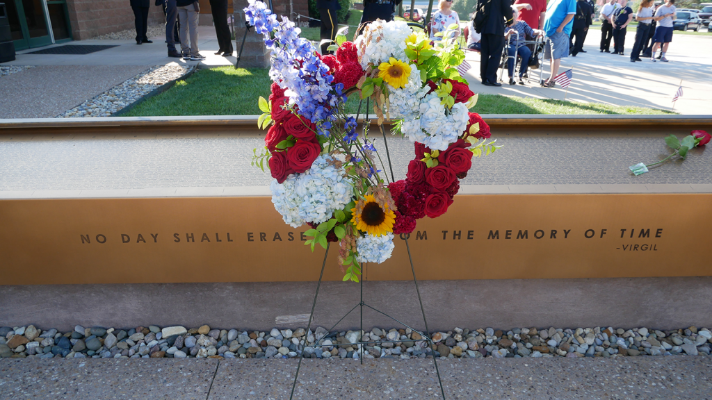 A flower wreath on a stand is placed at the 9/11 Memorial. Behind the wreath is the quote "No day shall erase you from the memory of time," attributed to Virgil.