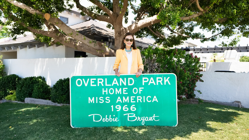 Debbie Bryant holds a large green and white street sign that reads "Overland Park; Home of Miss America 1966 Debbie Bryant."