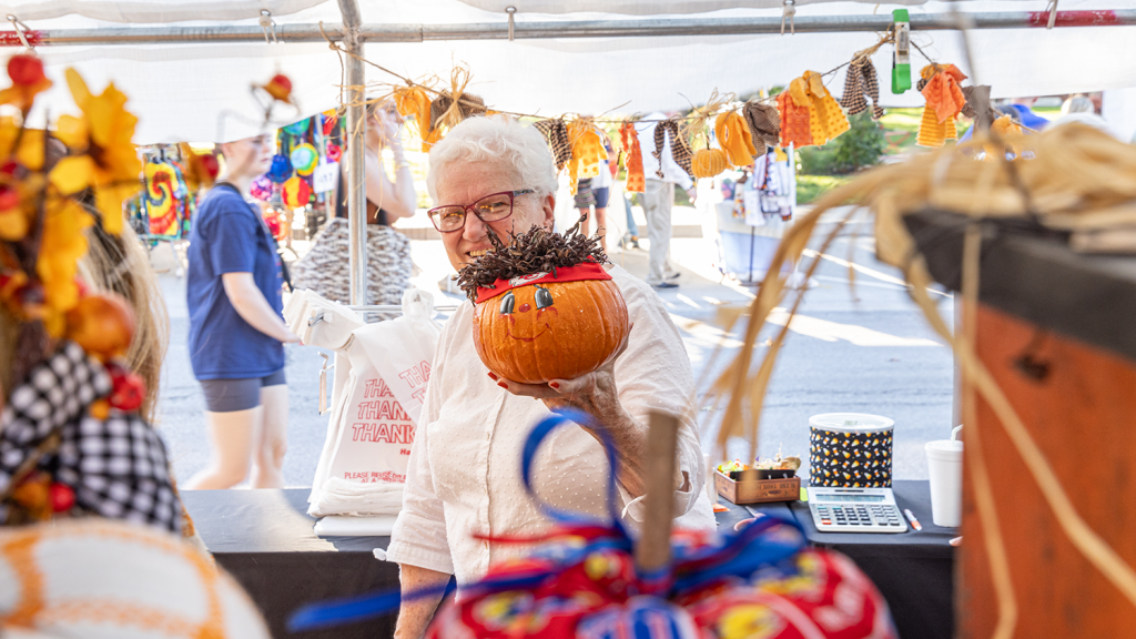 A woman holds a pumpkin with a painted face. On top of the pumpkin is a Chiefs sweatband and hair made of brown yarn, making the pumpkin resemble Chiefs player Patrick Mahomes.