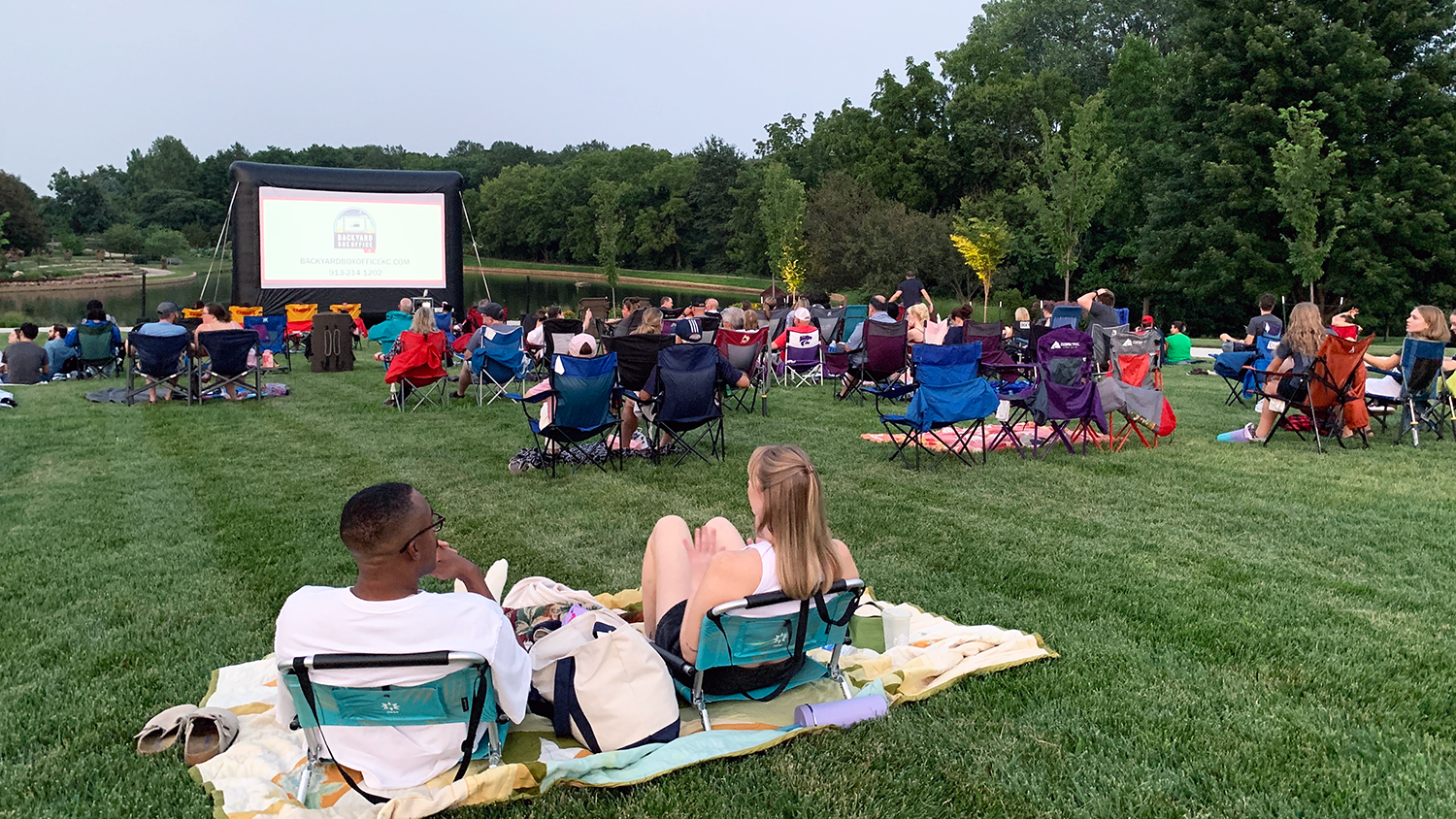 A crowd sits in lawn chairs or on picnic blankets outside, watching a movie on an inflatable movie screen.