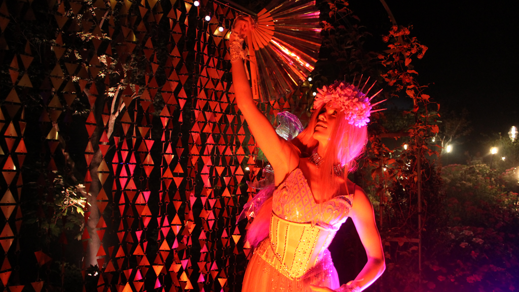A dancer holds a large fan at a performance, illuminated by red lights.