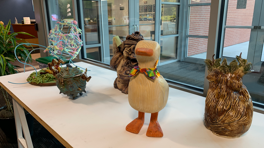 Various sculptures, one being a wooden duck wearing a bright bow tie, sit on a display table.