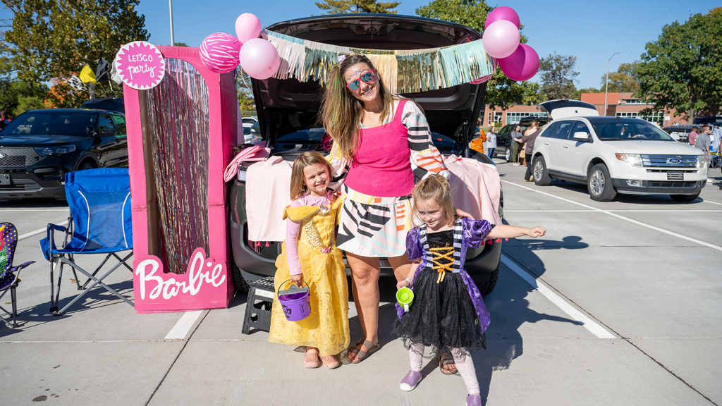 A woman and two young girls, all in Halloween costumes, smile outside a decorated car trunk.