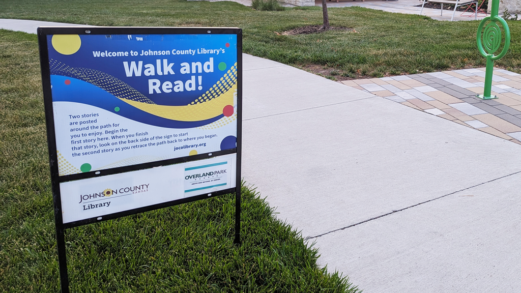A sign that reads "Welcome to Johnson County Library's Walk and Read" stands next to a park sidewalk.
