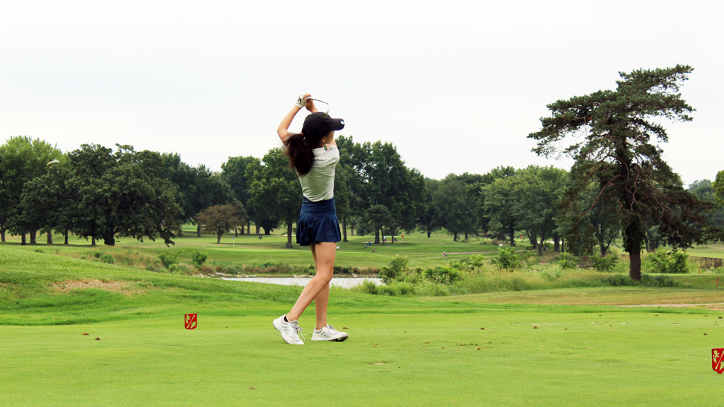 A woman on a golf course finishes her drive swing, holding her club above her head.