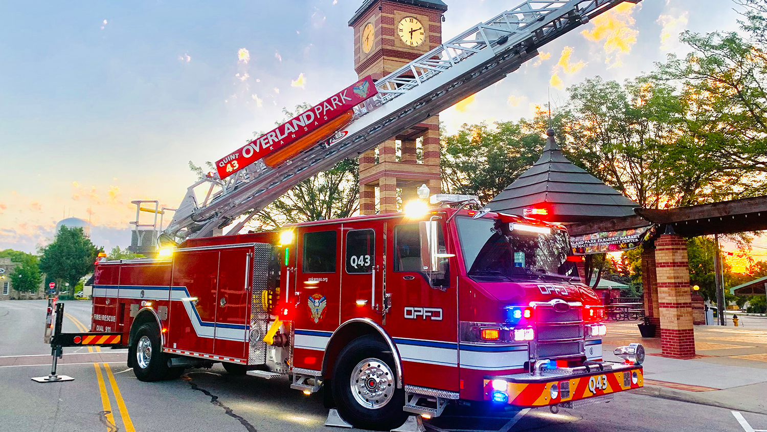 A firetruck parked in front of the Downtown Overland Park Clock Tower. The truck's red, blue and yellow lights are on, and the aerial ladder extends into the air.