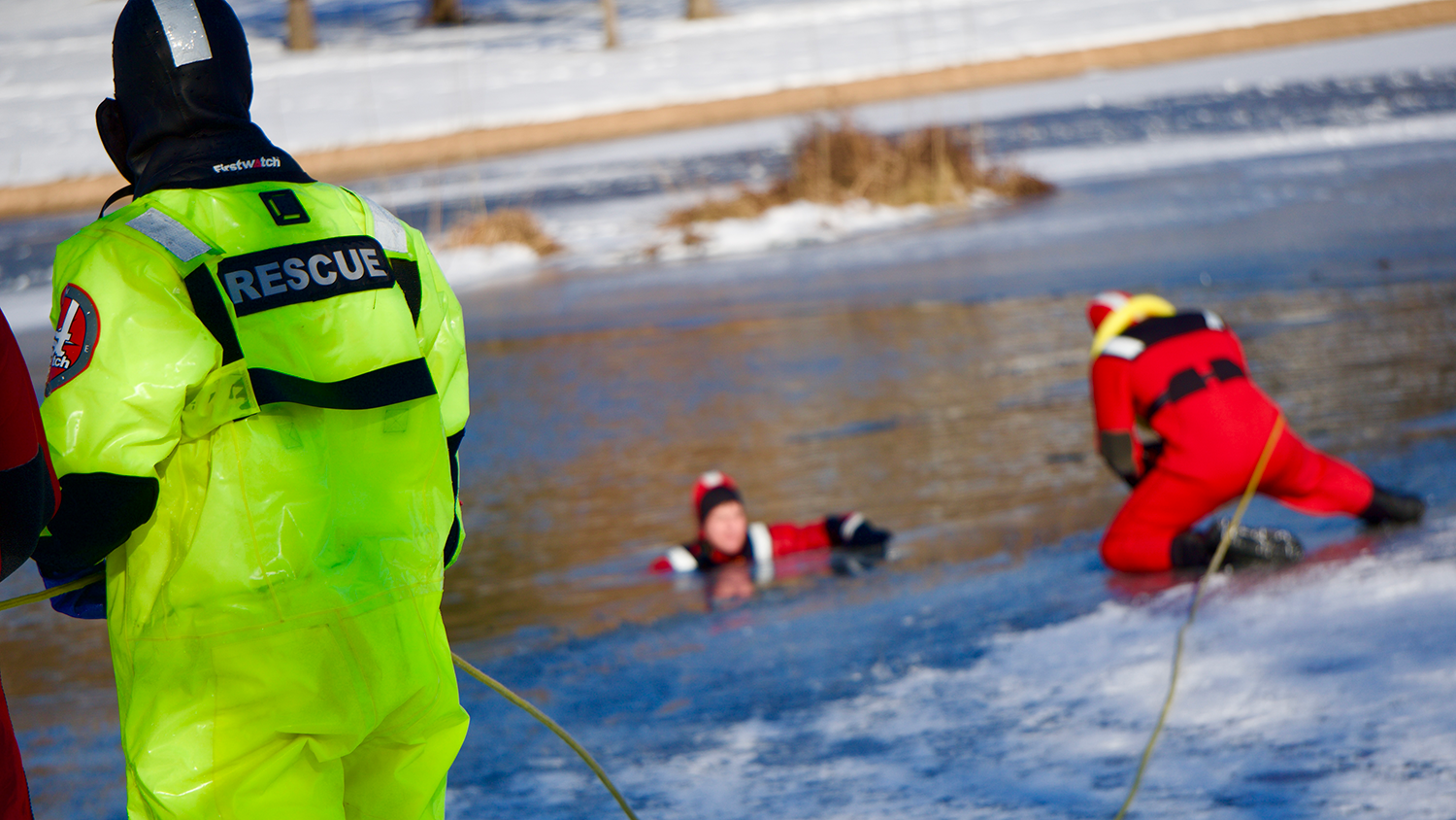 Three people complete Overland Park Fire Department Ice Training. During the training, one person swims in an icy lake, on person kneels down on the bank nearby with a rope, and one stands off to the side with the other end of the rope.