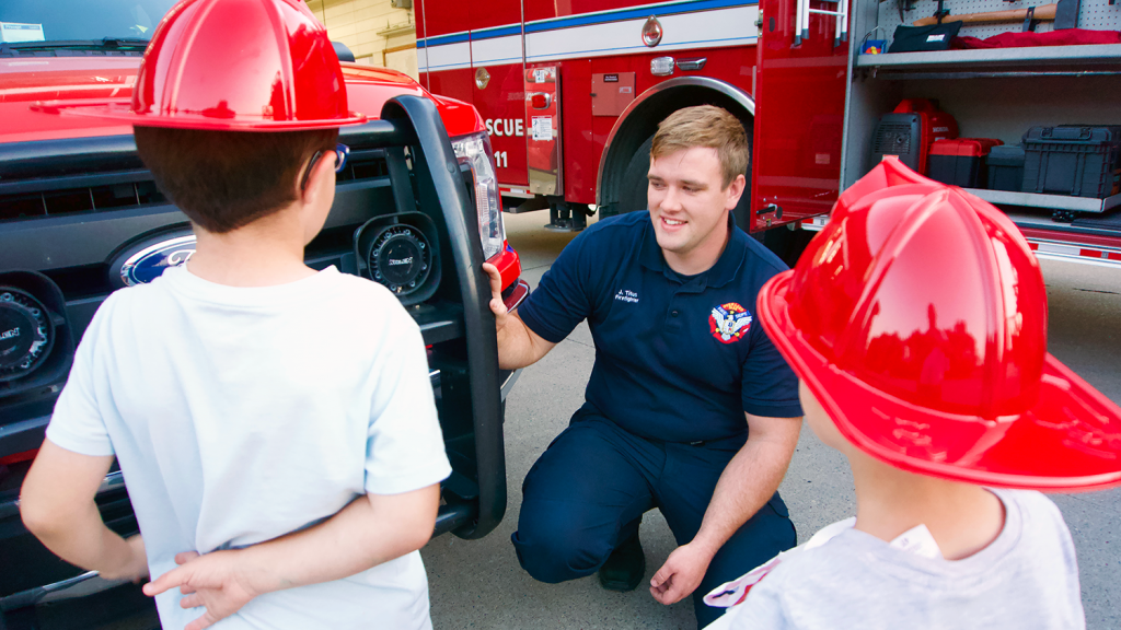 A firefighter kneels as he talks to two young boys, each wearing red, plastic firefighter helmets.