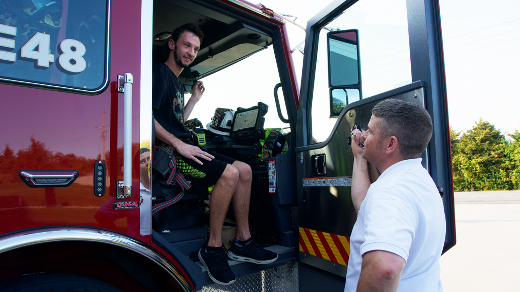 One man sits in the passenger-side seat of a fire truck, while a firefighter stands on the ground talking with him.