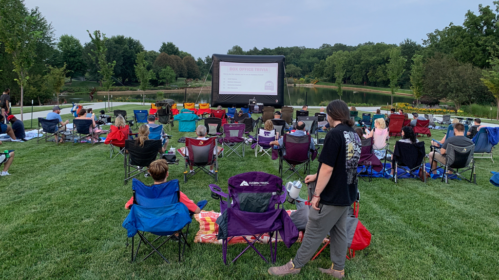 A crowd of people sit in lawn chairs watching a blow-up movie screen at the Arboretum.