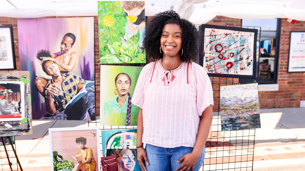 A woman stands smiling in front of her art display, showcasing various paintings of people.