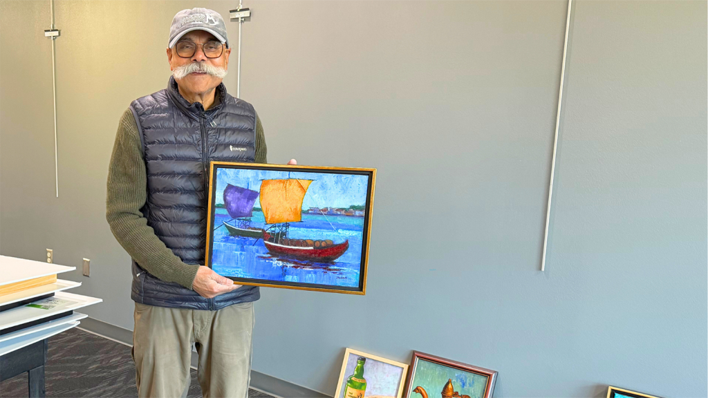 Dr. Jhulan Mukharji smiles, holding his painting of two boats on water.