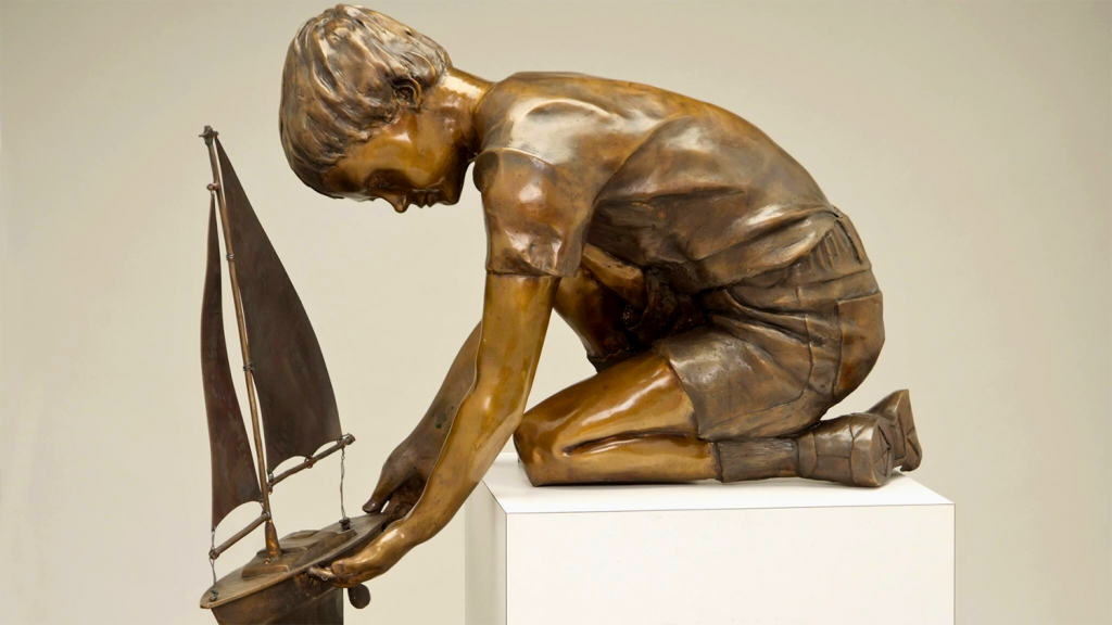 A bronze statue of a little boy lowering a model sail boat.
