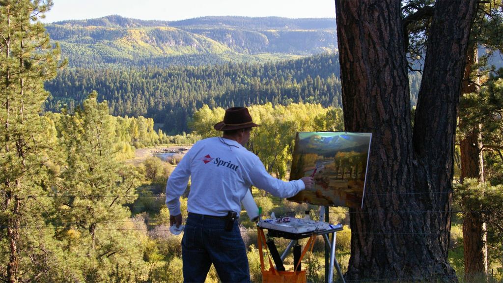 A man paints the landscape in front of him, which features small mountains and a forest.