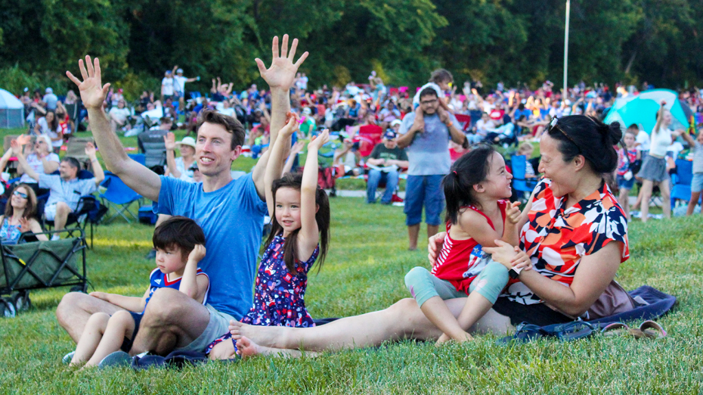 A family sits on a blanket in a grassy field with their arms raised, smiling.