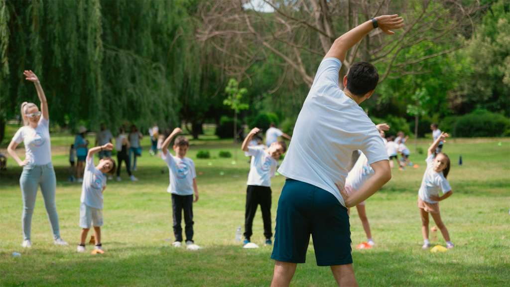 A man leads a group of young kids in a side stretch outside in a park.