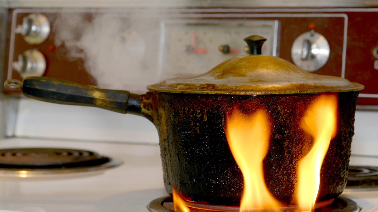 A pot with a lid sits on a gas stove, with an orange flame rising up its sides.