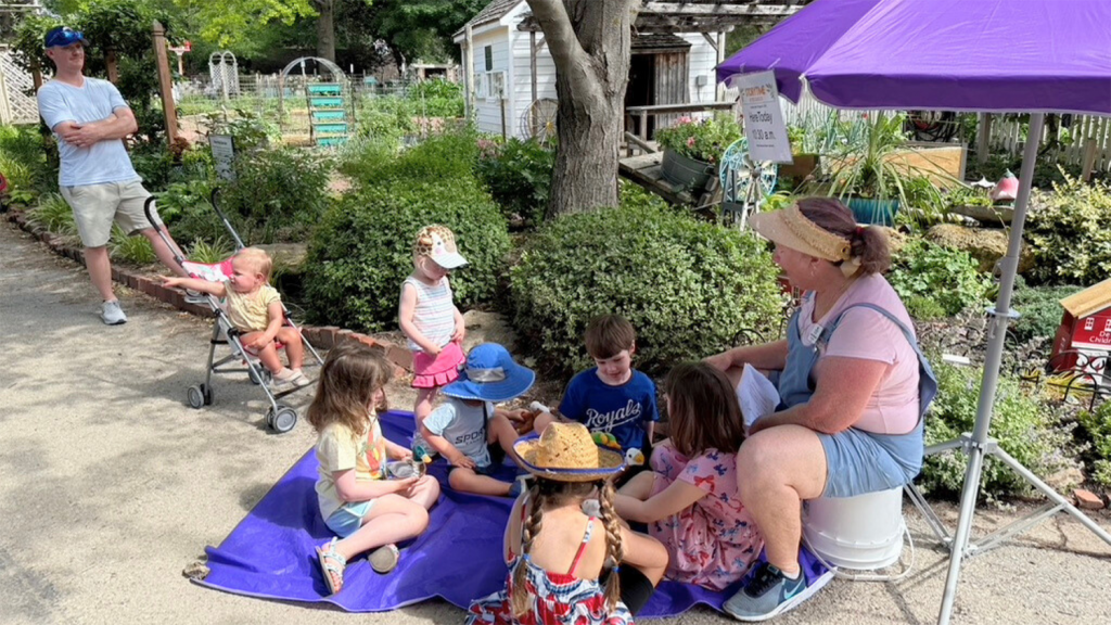 Kids sit outside on a purple picnic blanket listening to a woman tell a story.