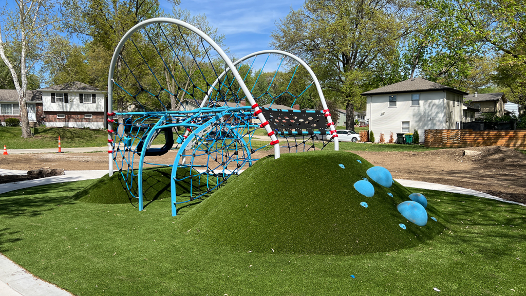 A piece of playground equipment spans between two turf hills. The bars of the equipment are connected with overlapping rope to climb on.