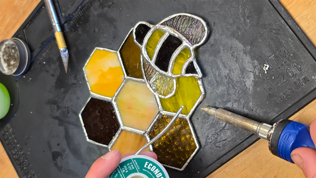 Two hands work with tools to create a piece of stained glass that features a bee and seven honeycomb hexagons.