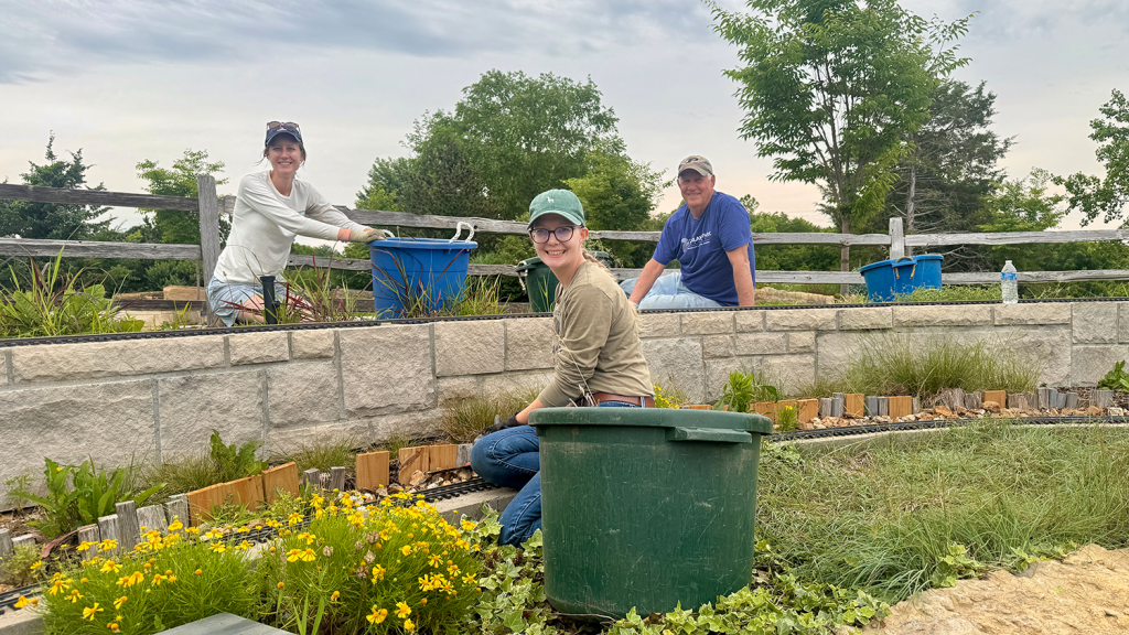 Three volunteers tend to plants in the Train Garden at the Overland Park Arboretum & Botanical Gardens. Each volunteer wears a hat and gloves, and work next to large circular bins.