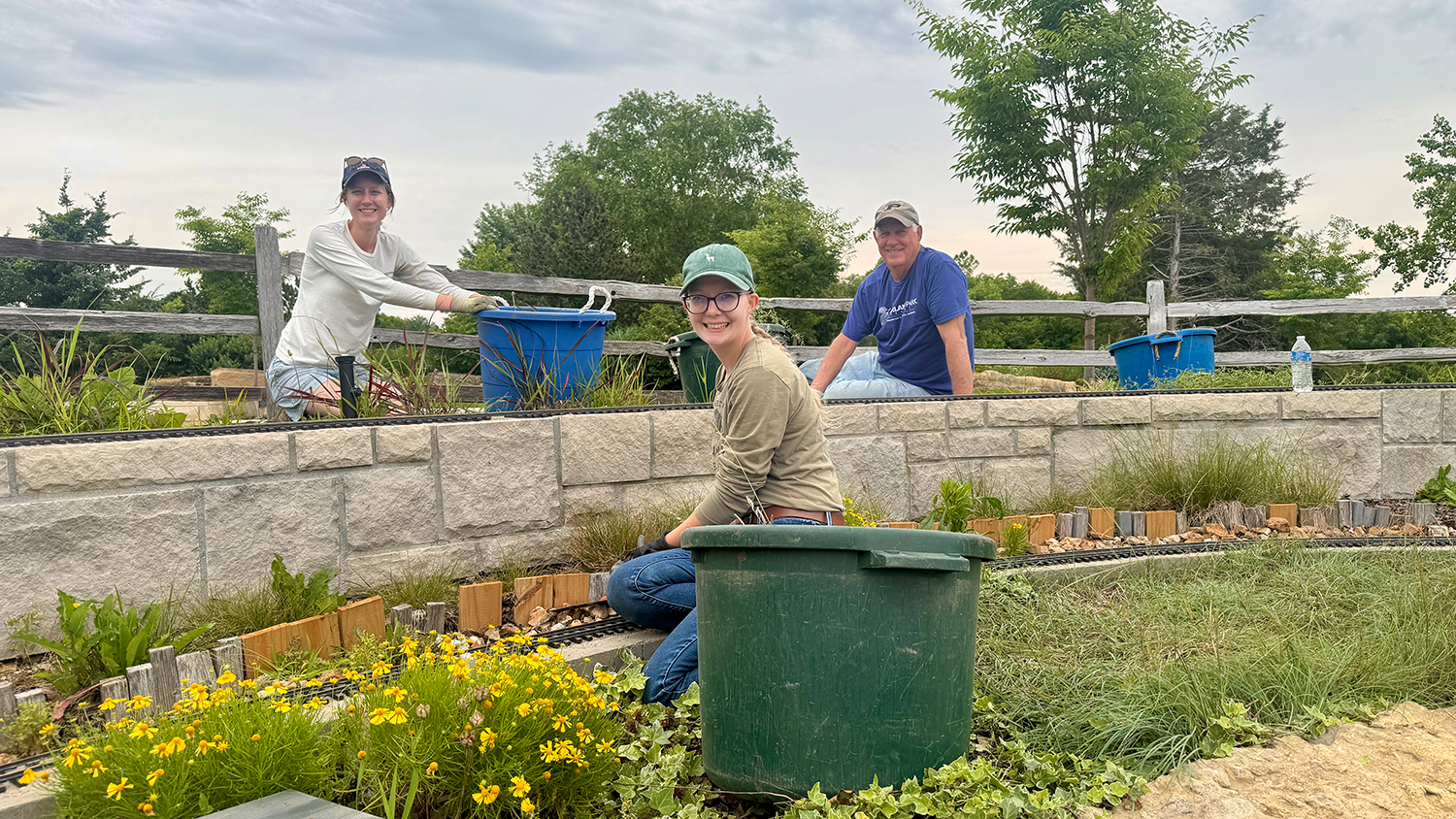Three volunteers tend to plants in the Train Garden at the Overland Park Arboretum & Botanical Gardens. Each volunteer wears a hat and gloves, and work next to large circular bins.