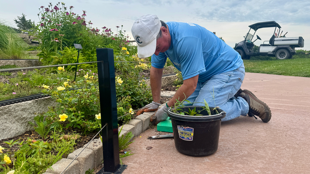 A volunteer repairs a row of stones along a garden path at the Overland Park Arboretum & Botanical Gardens. Next to him sits a black bin of pulled weeds.