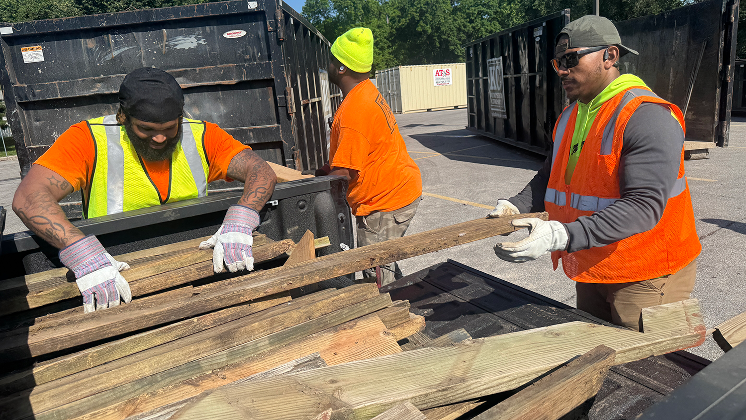 Two volunteers unload long wooden construction debris from the bed of a truck. Both of the volunteers wear bright construction vests and thick gloves.