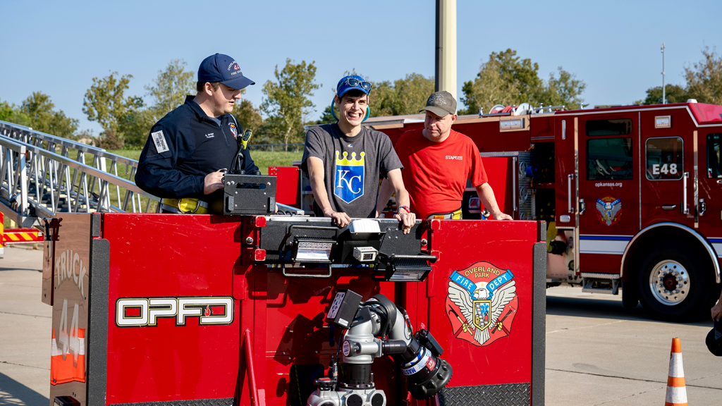 Two Overland Park firefighters stand in the lowered basket platform of a firetruck ladder at Friends of First Responders Day. Standing between them is a man, smiling and holding onto the sides of the basket.